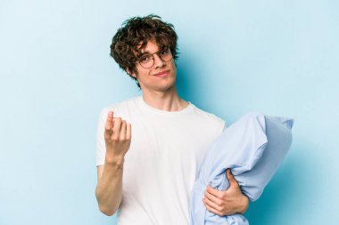 Genç beyaz adam pijama giyiyor ve yastığı mavi arka planda izole halde tutuyor. Sanki seni davet ediyormuş gibi..