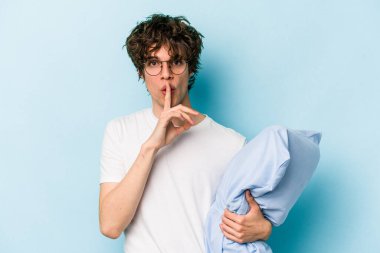 Genç, beyaz bir adam pijama giyiyor ve mavi arka planda yastık tutuyor. Sır saklıyor ya da sessizlik istiyor..