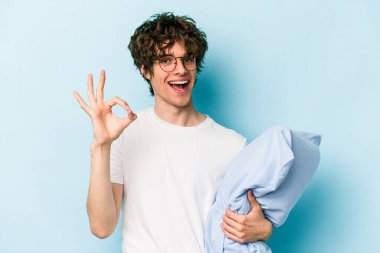 Genç beyaz adam pijama giyiyor ve yastık tutuyor. Mavi arka planda izole edilmiş. Neşeli ve kendinden emin..