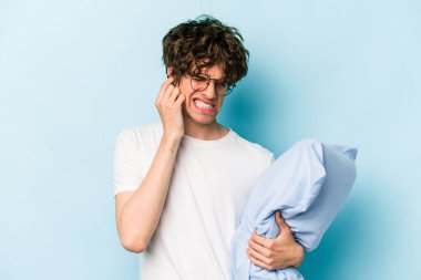 Pijama giyen ve yastık tutan genç beyaz adam mavi arka planda izole edilmiş kulakları elleriyle kapatıyor..