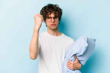 Genç beyaz bir adam pijama giyiyor ve yastığı mavi arka planda tutuyor. Kameraya yumruğunu gösteriyor, agresif yüz ifadesi var..