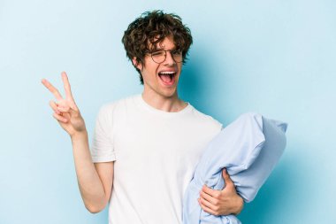 Pijama giyen ve yastık tutan genç beyaz adam mavi arka planda izole edilmiş neşeli ve kaygısız parmaklarıyla barış sembolü gösteriyor..