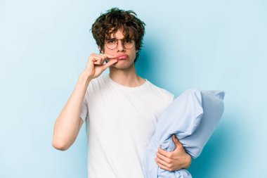 Genç, beyaz bir adam pijama giyiyor ve yastığı mavi arka planda, parmakları dudaklarında gizli tutuyor..