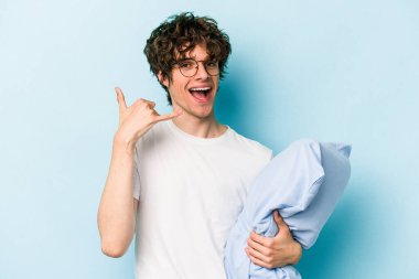 Genç beyaz bir adam pijama giyiyor ve yastık tutuyor. Mavi arka planda izole edilmiş. Cep telefonu hareketini parmaklarıyla gösteriyor..