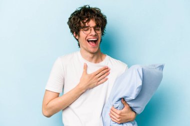 Genç beyaz bir adam pijama giyiyor ve mavi arka planda yastık tutuyor. Gülüyor, elini göğsüne koyuyor..