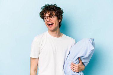 Genç beyaz adam pijama giyiyor ve yastığı mavi arka planda yalnız tutuyor. Gülümsüyor, neşeli ve keyifli görünüyor..