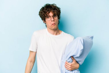 Genç beyaz adam pijama giyiyor ve yastık tutuyor. Mavi arka planda izole edilmiş. Omuzlarını silkiyor ve gözleri açık..