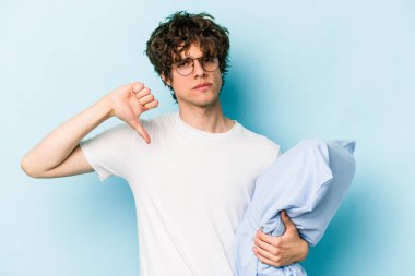 Genç beyaz bir adam pijama giyiyor ve yastığı mavi arka planda izole halde tutuyor. Sevimsiz bir jest yapıyor. Anlaşmazlık konsepti.
