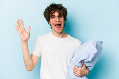 Genç beyaz adam pijama giyiyor ve yastığı mavi arka planda yalnız tutuyor. Hoş bir sürprizle karşılaşıyor, heyecanlı ve el kaldırıyor..