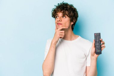 Elinde televizyon kumandası olan beyaz bir adam mavi arka planda izole edilmiş şüpheli ve şüpheci bir ifadeyle yana bakıyor..