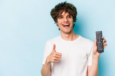 Elinde televizyon kumandası olan beyaz bir adam mavi arka planda izole edilmiş gülümsüyor ve başparmağını kaldırıyor.