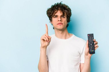 Elinde televizyon kumandası olan beyaz bir adam mavi arka planda bir numarayı parmağıyla gösteriyor..