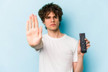 Elinde televizyon kumandası olan beyaz bir adam, mavi arka planda izole edilmiş, elini uzatmış, dur işaretini göstererek, seni engelliyor..
