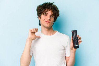Mavi arka planda izole edilmiş televizyon kumandası tutan genç beyaz adam gururlu ve kendinden emin hissediyor, örnek olarak.