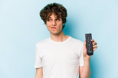 Elinde televizyon kumandası olan beyaz bir adam mavi arka planda izole edilmiş, kafası karışmış, şüpheli ve güvensiz hissediyor..