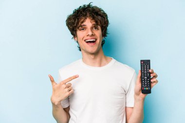 Elinde televizyon kumandası olan beyaz bir adam, mavi arka planda izole edilmiş, gömlek fotokopi alanını işaret ediyor, gururlu ve kendinden emin.