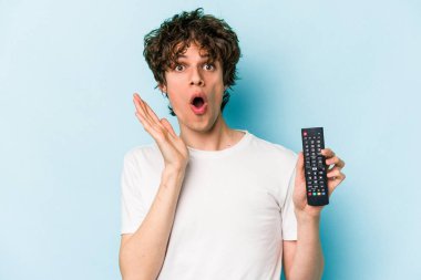 Elinde televizyon kumandası olan beyaz bir adam mavi arka planda izole edilmiş. Şaşırmış ve şok olmuş..