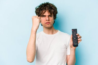 Elinde televizyon kumandası olan genç beyaz bir adam mavi arka planda izole edilmiş kameraya yumruk gösteriyor, agresif bir yüz ifadesi var..