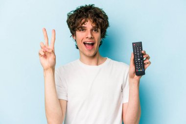 Elinde televizyon kumandası olan beyaz bir adam mavi arka planda iki numarayı parmaklarıyla gösteriyor..