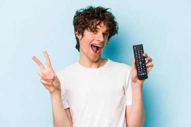 Elinde televizyon kumandası olan beyaz bir adam mavi arka planda izole edilmiş neşeli ve kaygısız parmakları olan bir barış sembolü gösteriyor..