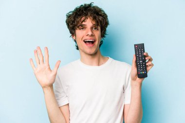 Elinde televizyon kumandası olan beyaz bir adam mavi arka planda izole edilmiş gülümseyen beş numarayı parmaklarıyla gösteriyor..