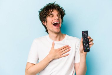 Mavi arka planda izole edilmiş televizyon kumandası tutan genç beyaz adam kahkahalar atıyor..