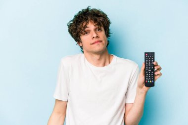 Elinde televizyon kumandası olan beyaz bir genç mavi arka planda izole edilmiş omuzlarını silkeliyor ve gözleri açık..