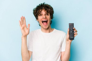 Elinde televizyon kumandası olan beyaz bir adam mavi arka planda izole edilmiş. Hoş bir sürprizle karşılaşıyor, heyecanlı ve el kaldırıyor..