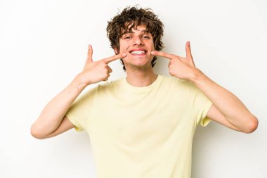 Beyaz arka planda izole edilmiş genç beyaz adam gülümsüyor, parmaklarını ağıza doğrultuyor..