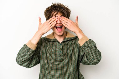 Beyaz arka planda soyutlanmış beyaz tenli genç bir adam elleriyle gözlerini kapatıyor, geniş bir gülümsemeyle bir sürpriz bekliyor..