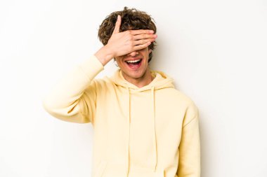 Beyaz arka planda soyutlanmış beyaz tenli genç bir adam elleriyle gözlerini kapatıyor, geniş bir gülümsemeyle bir sürpriz bekliyor..