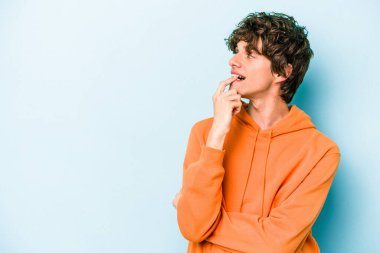 Mavi arka planda izole edilmiş genç beyaz adam şüpheli ve şüpheci bir ifadeyle yana bakıyor..