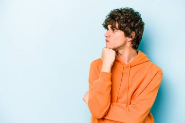 Mavi arka planda izole edilmiş genç beyaz adam şüpheli ve şüpheci bir ifadeyle yana bakıyor..