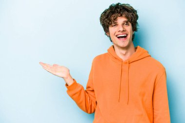 Mavi arka planda izole edilmiş genç beyaz bir adam bir palmiye üzerinde fotokopi alanı ve belinde bir el daha gösteriyor..