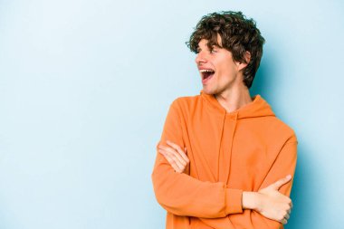 Mavi arka planda izole edilmiş genç beyaz adam kollarını kavuşturarak gülümsüyor..