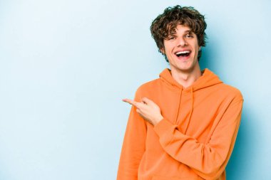 Mavi arka planda izole edilmiş genç beyaz adam işaret parmağıyla işaret ederek gülümsüyor..