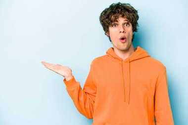 Mavi arka planda izole edilmiş genç beyaz adam, avuç içi fotokopi alanını elinde tutmaktan etkilenmiş..