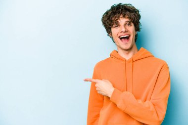 Mavi arka planda izole edilmiş genç beyaz adam gülümsüyor ve boş alanda bir şeyler gösteriyor..