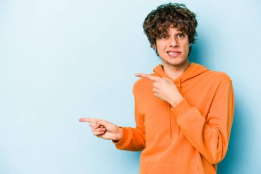 Mavi arka planda izole edilmiş genç beyaz adam işaret parmağıyla fotokopi alanını işaret ederek şok oldu..