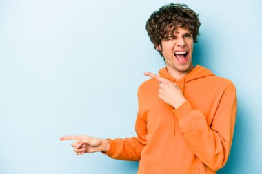 Mavi arka planda izole edilmiş genç beyaz adam işaret parmağıyla fotokopi alanını işaret ederek heyecan ve arzusunu ifade ediyor..