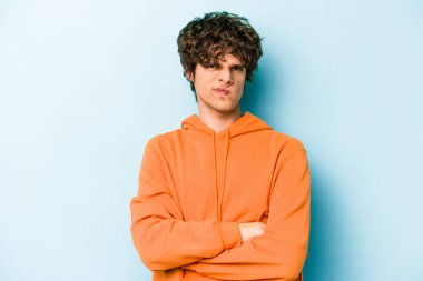 Kafkasyalı genç adam mavi arka planda izole edilmiş hoşnutsuzca kaşlarını çatmış, kollarını kavuşturmuş..