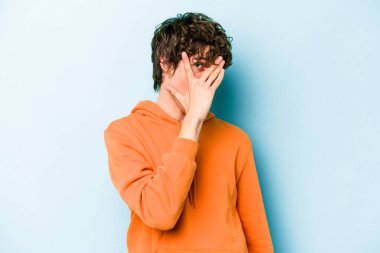 Mavi arka planda izole edilmiş genç beyaz adam kameraya parmaklarıyla göz kırpıyor, yüzünü kapatmaktan utanıyor..