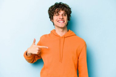 Mavi arka planda izole edilmiş genç beyaz adam bir gömlek fotokopi alanını işaret ediyor, gururlu ve kendinden emin.