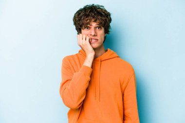 Mavi arka planda izole edilmiş, tırnak yiyen, gergin ve endişeli genç bir beyaz adam..