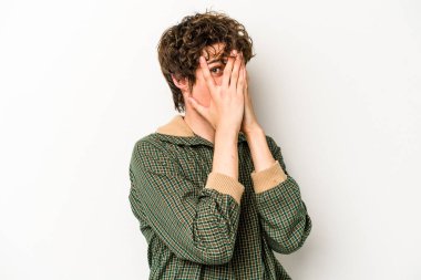 Beyaz arka planda izole edilmiş genç beyaz bir adam parmaklarının arasından korkmuş ve gergin bir şekilde göz kırpıyor..