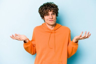 Kafkasyalı genç adam mavi arka planda izole edilmiş sorgulama jestinde şüphe ediyor ve omuz silkiyor..