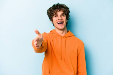 Mavi arka planda izole edilmiş genç beyaz adam selamlama jesti olarak kameraya elini uzatıyor..