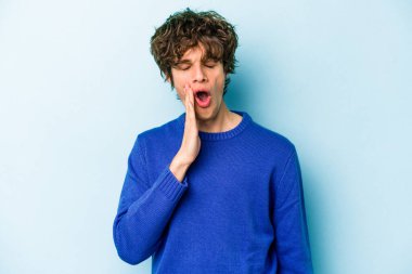 Mavi arka planda izole edilmiş genç beyaz adam esniyor. Ağzı eliyle kapatan yorgun bir hareket sergiliyor..