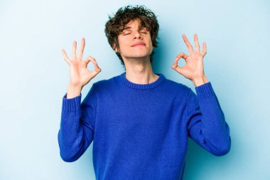 Mavi arka planda izole edilmiş genç beyaz adam yorucu bir günün ardından rahatlıyor. Yoga yapıyor..