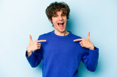 Mavi arka planda izole edilmiş genç beyaz adam parmağını göstererek şaşırdı, geniş bir gülümsemeyle..